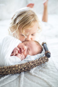 newborn on bed in basket big sister kissing his forehead